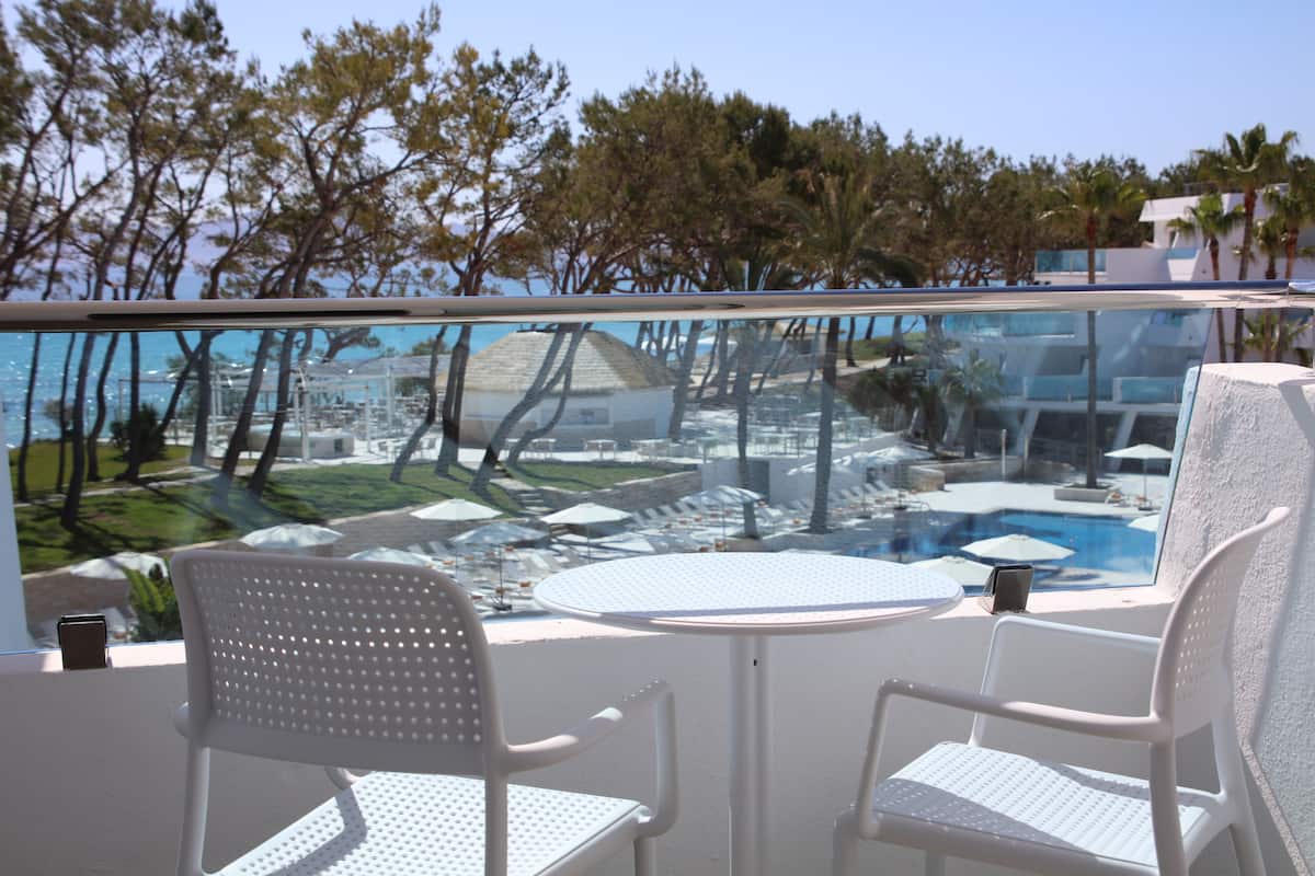 Ein Balkon mit zwei weißen Stühlen und einem Tisch, mit Blick auf einen Poolbereich mit Sonnenschirmen und Palmen.