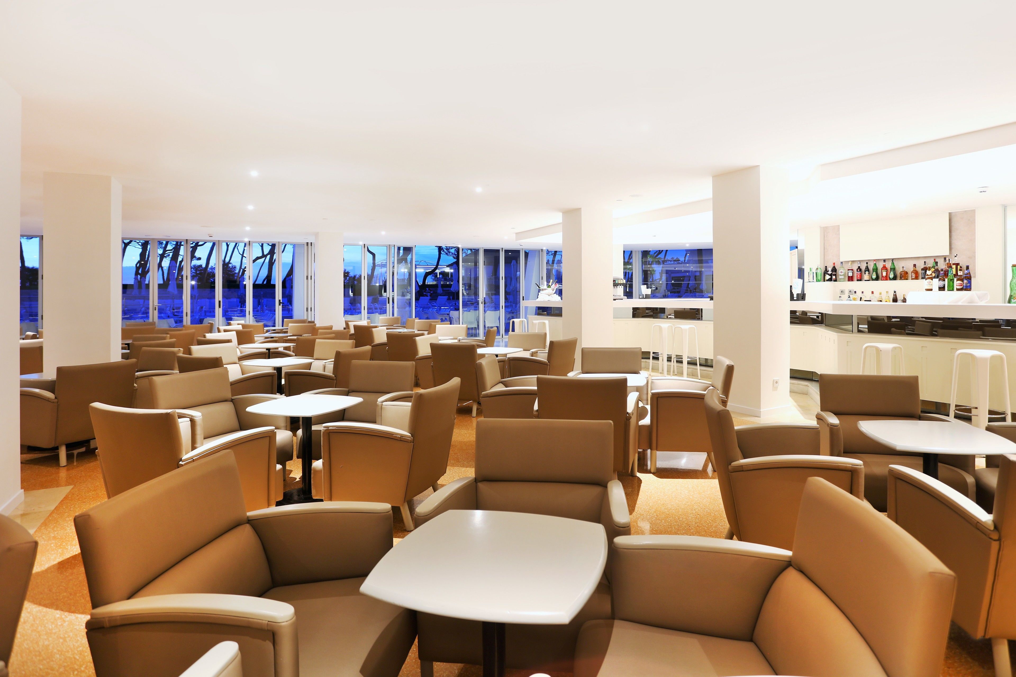 A spacious indoor seating area with multiple tables and chairs, large windows offering a view of the outside, and a bar with a variety of bottles.