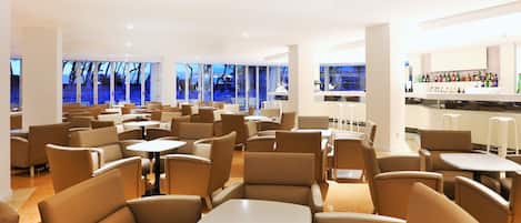 A spacious indoor seating area with multiple tables and chairs, large windows offering a view of the outside, and a bar with a variety of bottles.