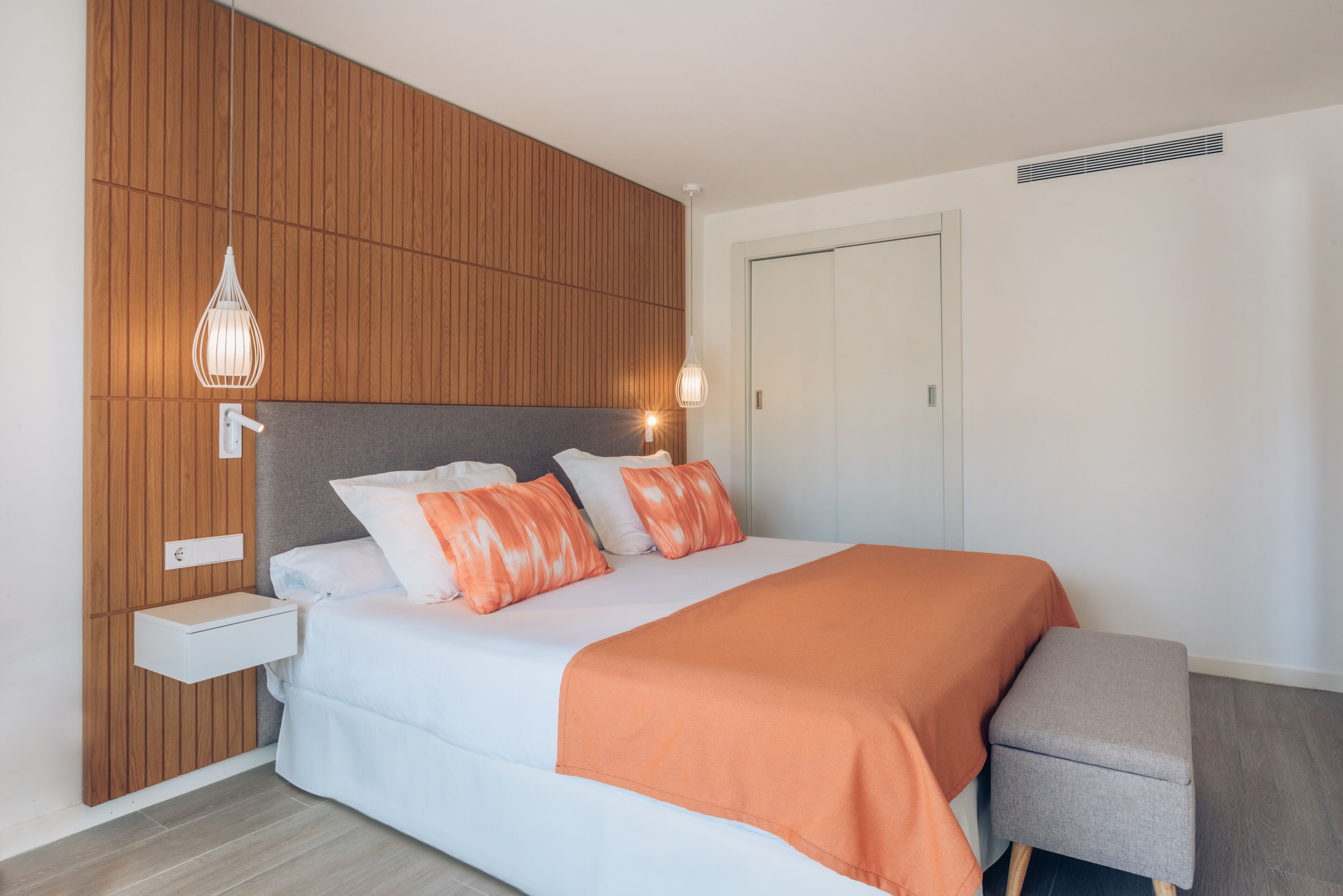 A modern bedroom with a wooden panel wall, a bed with white and orange bedding, a bedside table, and a bench.