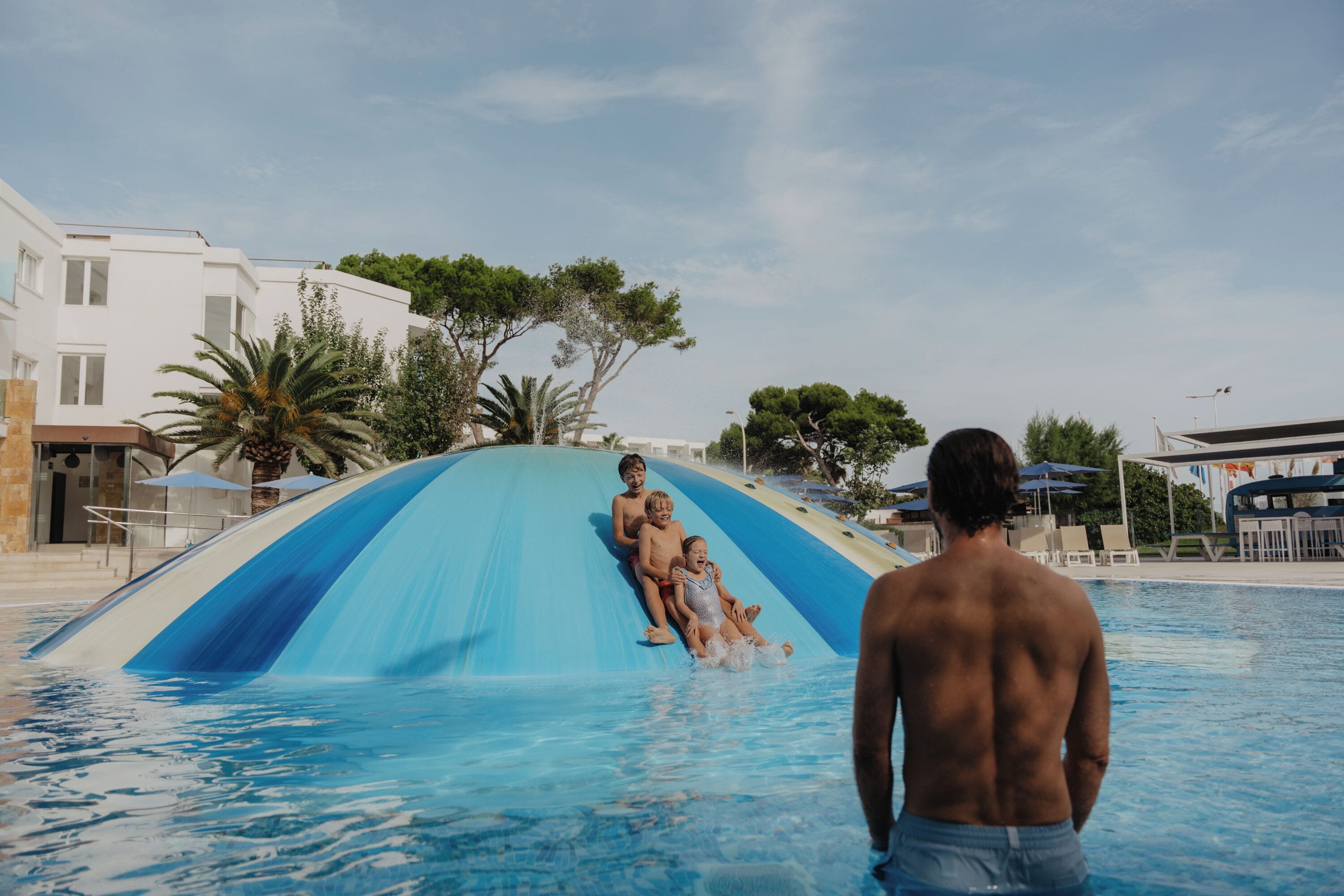A swimming pool with a blue and white water slide, surrounded by buildings and palm trees.