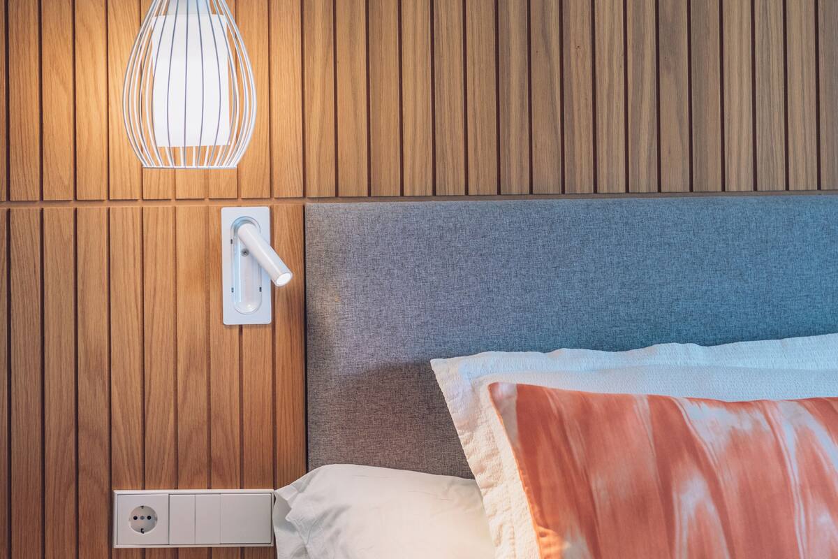 Ein ordentlich bezogenes Bett mit grauem Kopfteil, einem weißen Nachttisch und einer wandmontierten Leuchte.