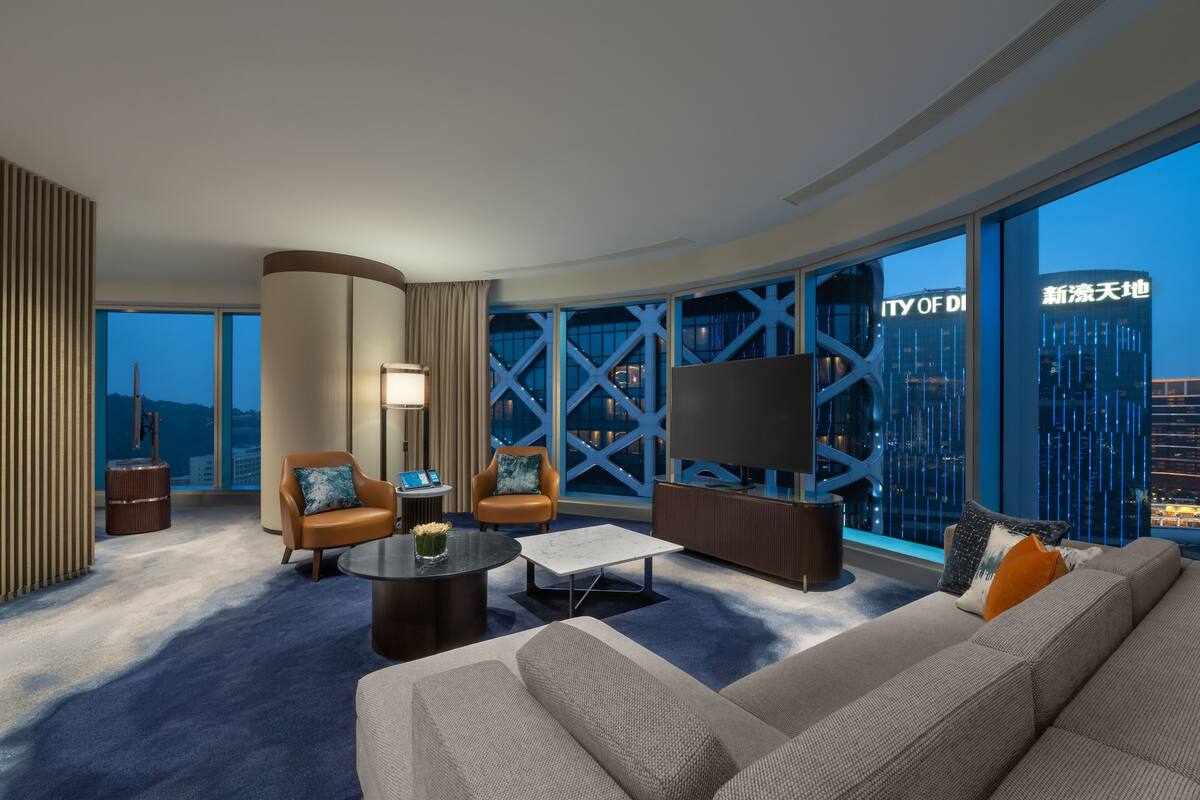 A modern hotel room with a large window, a sofa, a coffee table, and a TV.