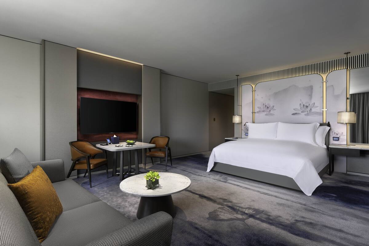 A modern hotel room with a large bed, a sofa, two chairs, a small table with a plant, and a TV.