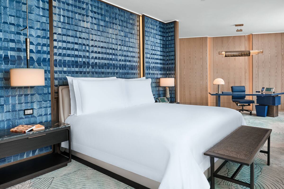 A hotel room with a large bed, two bedside tables, a desk with a chair, and a patterned wall.