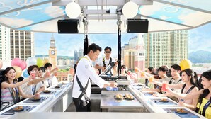 Un grupo de personas cenando en un restaurante al aire libre con vistas a la ciudad, incluyendo una torre de reloj, mientras un chef prepara comida.