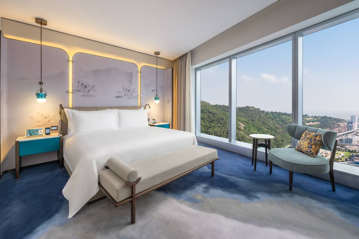 A modern hotel room with a large bed, a small table, a chair, and a view of a cityscape through large windows.