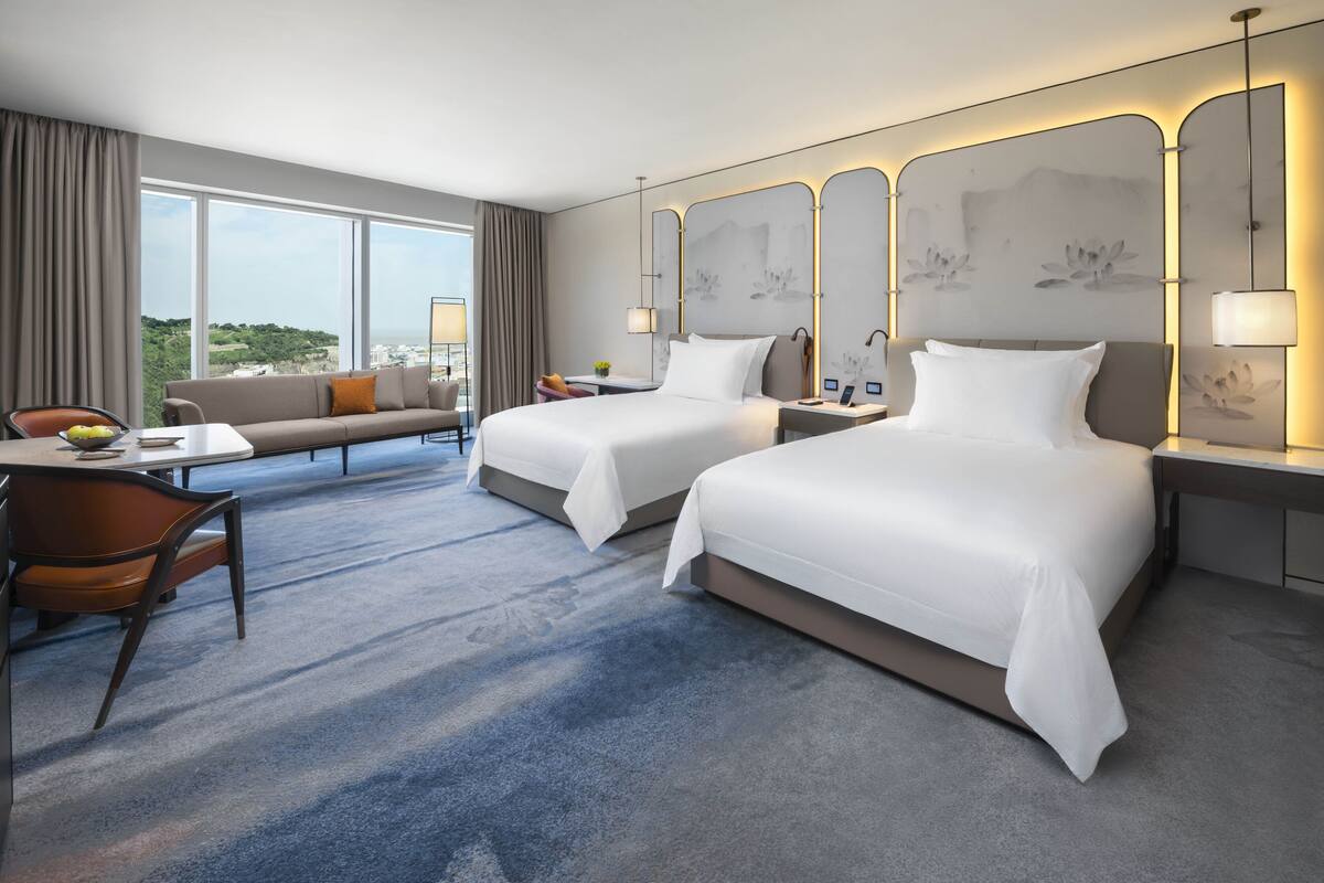 A hotel room with two beds, a sofa, a desk with a chair, and large windows offering a view of the city.