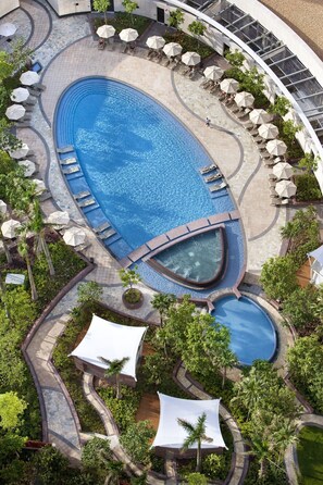 A large oval-shaped swimming pool with multiple lounge areas and umbrellas, surrounded by palm trees and a stone pathway. - City of Dreams – Nüwa Macau (Cotai)
