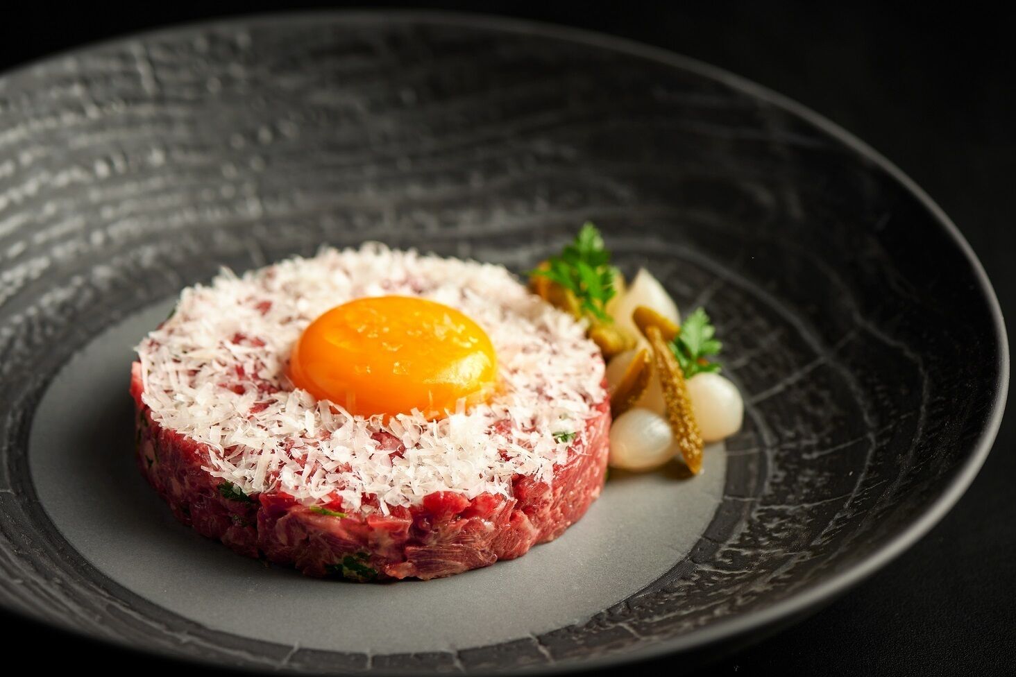 A dish of raw steak topped with shredded cheese and a raw egg yolk, garnished with herbs and pickles on a dark plate.