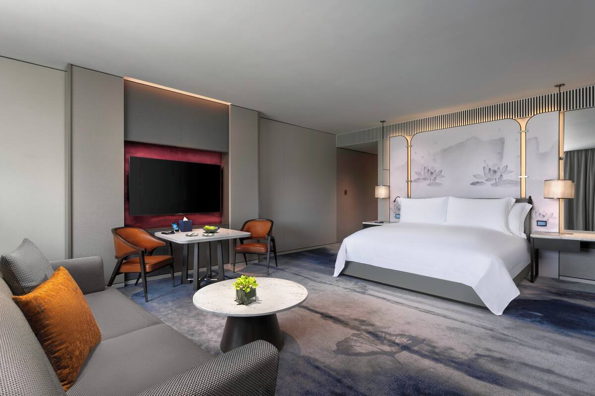 A modern hotel room with a large bed, a sofa, two chairs, a small table with a plant, and a TV.