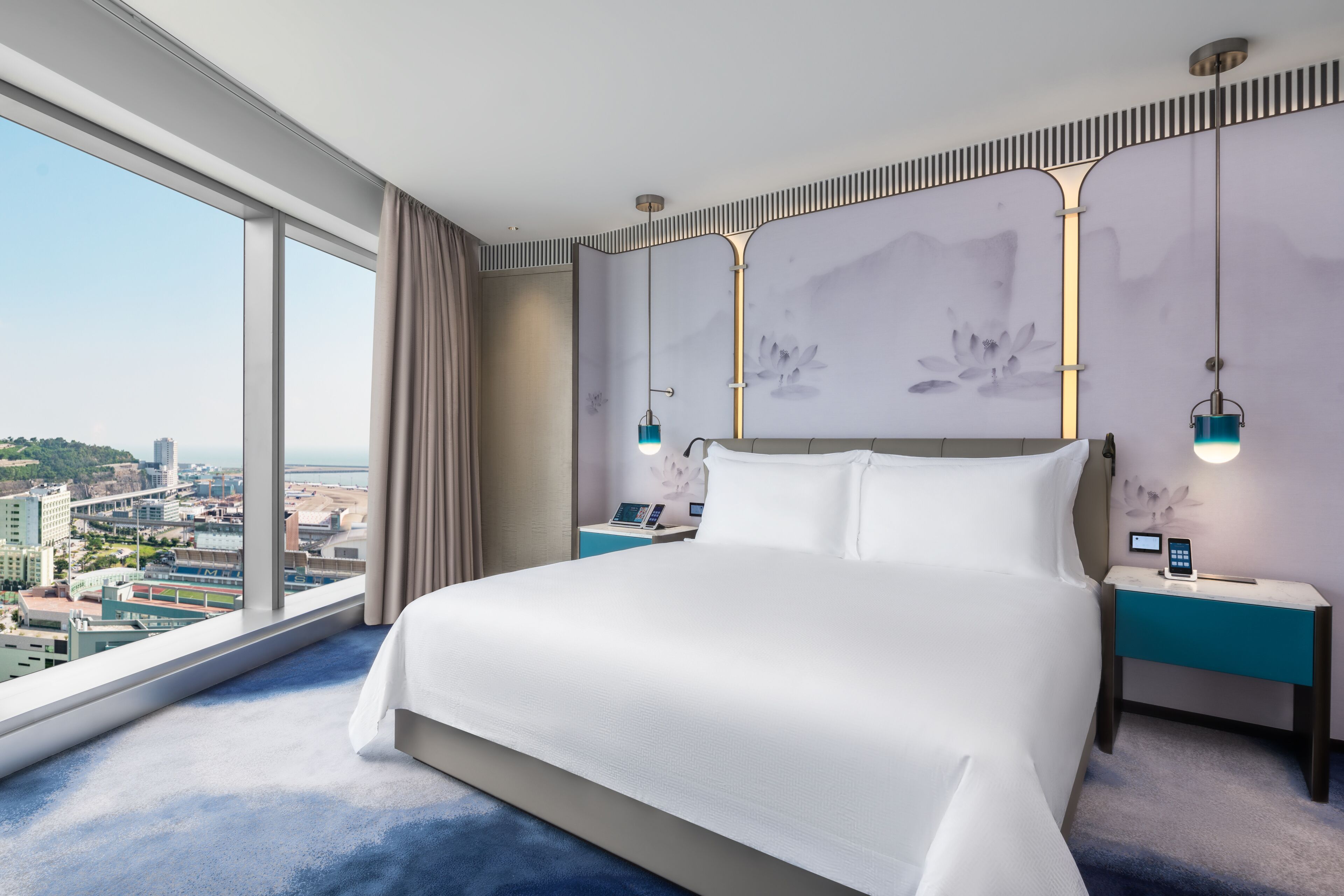 A modern hotel room with a large bed, a city view, and a desk with a phone.