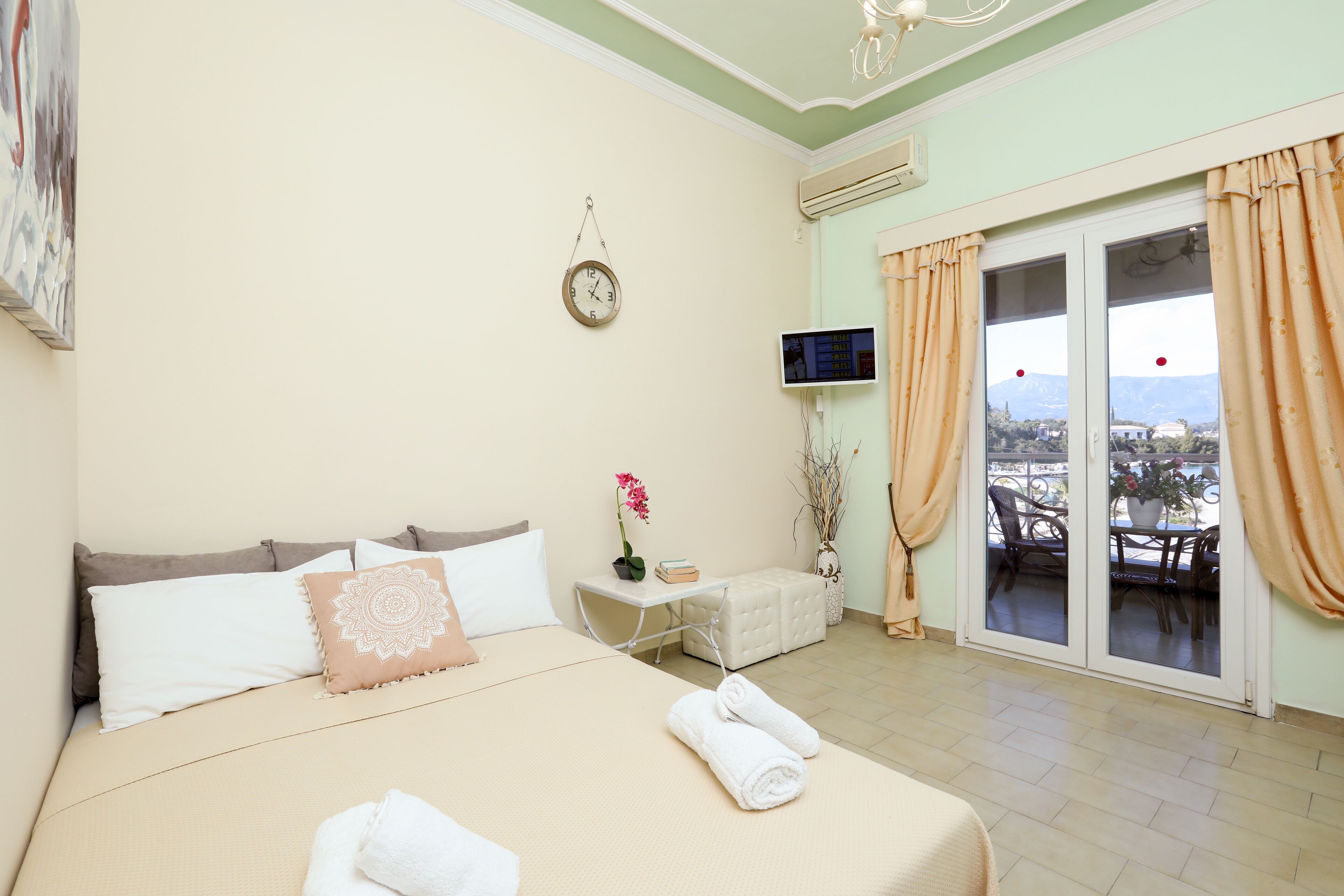 A hotel room with a bed, a TV, a window with curtains, a small table with a plant, and a view of the outside.