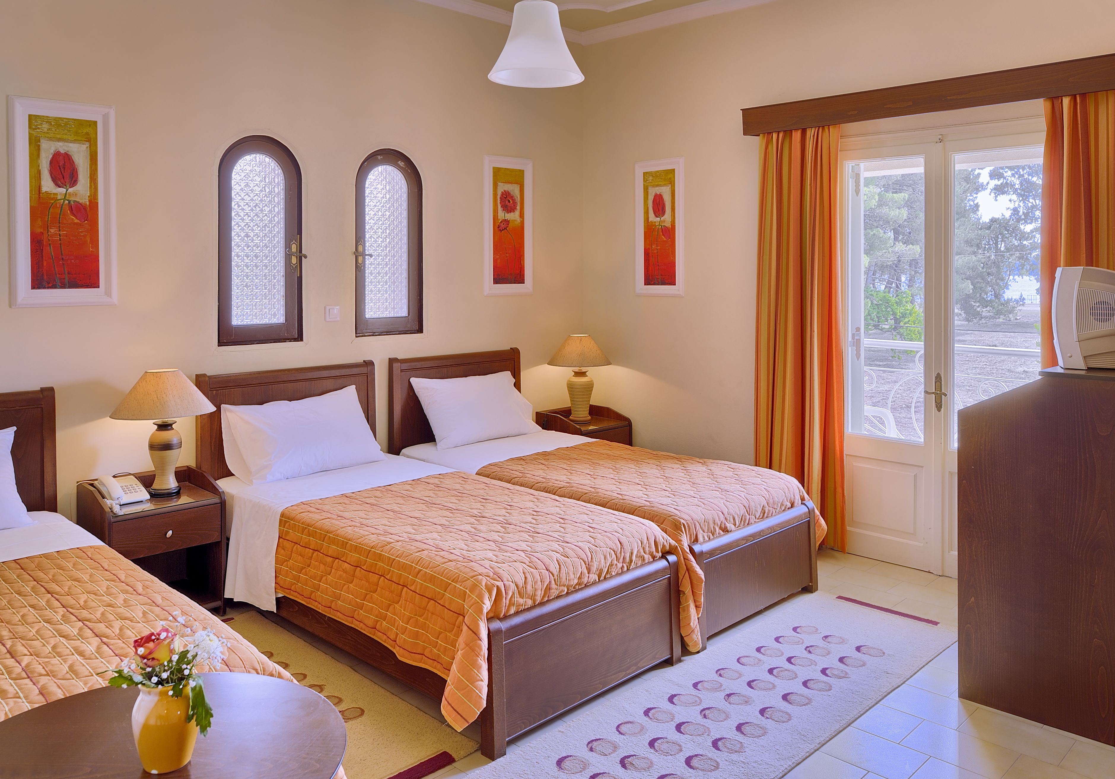 Habitación de hotel con dos camas, un escritorio, una silla, una lámpara, un florero con flores y vistas al exterior a través de una ventana.