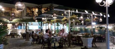 An outdoor dining area with tables and chairs, surrounded by potted plants and hanging lights.