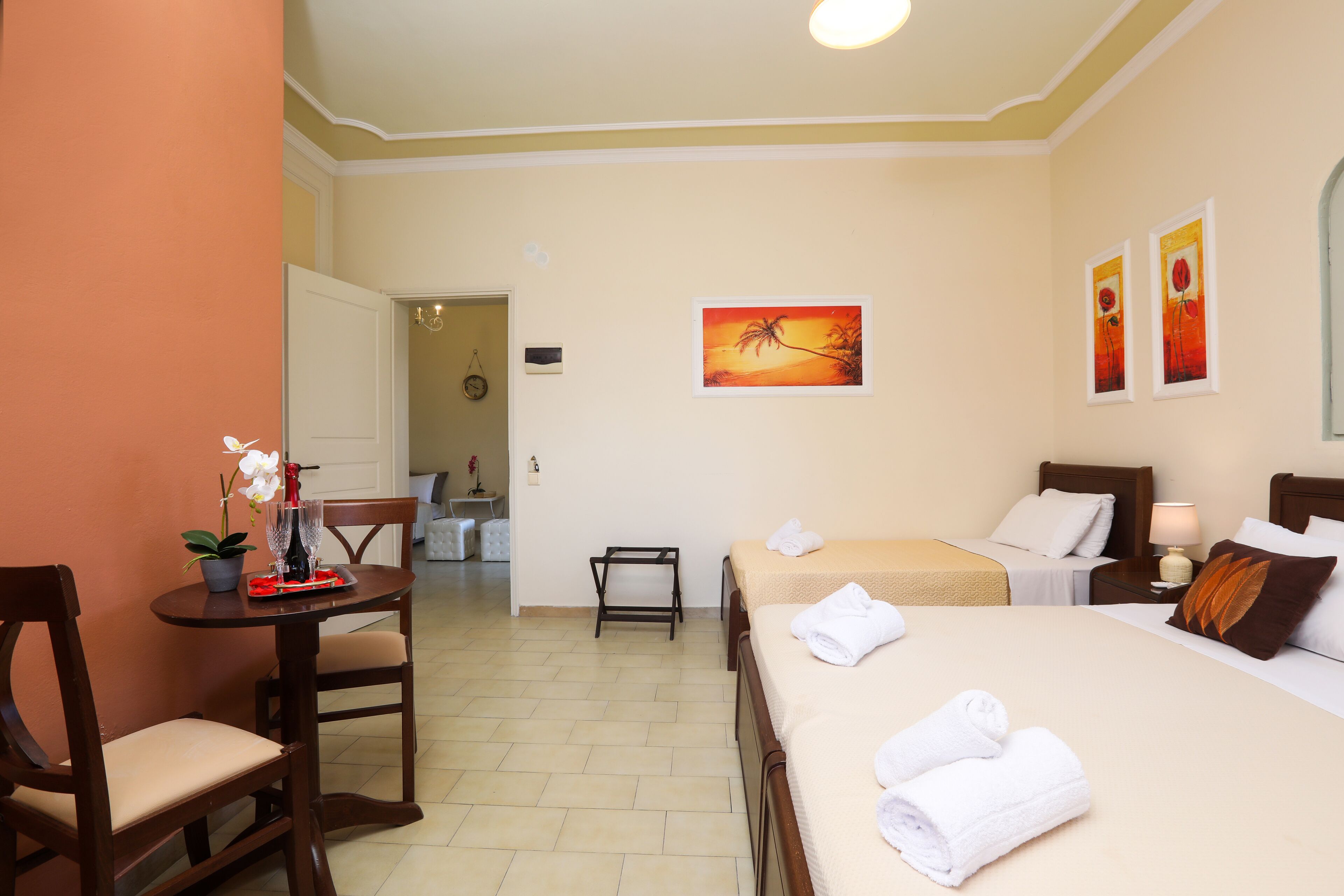 A hotel room with two beds, a dining table with chairs, a small table with a vase and flowers, and artwork on the walls.