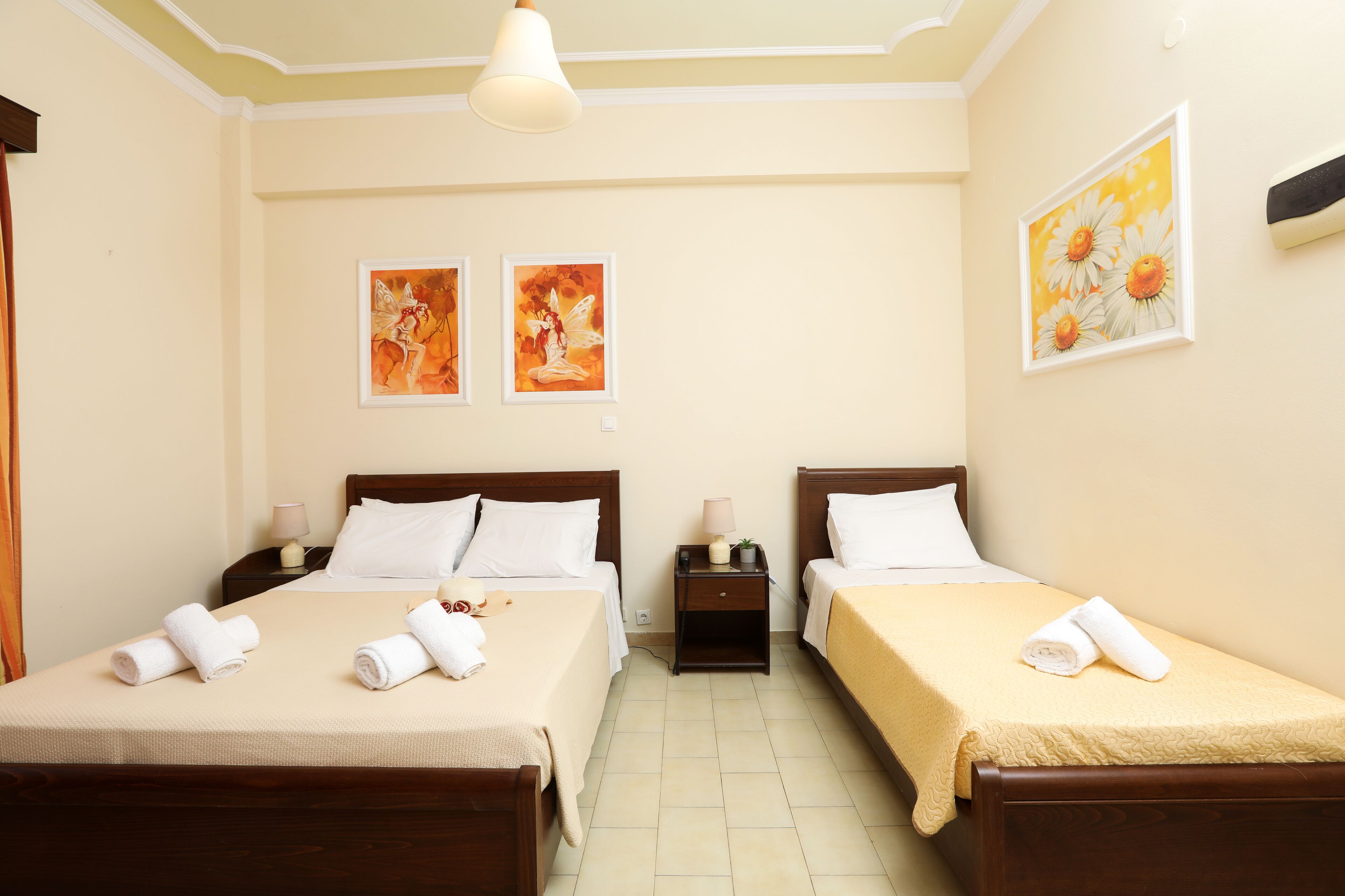 Une chambre d’hôtel avec deux lits, chacun doté d’une tête de lit en bois, de draps blancs et de serviettes. Il y a deux tables de chevet avec des lampes et une petite plante en pot. Les murs sont peints dans une couleur claire, et deux œuvres d’art encadrées sont accrochées au-dessus des lits.