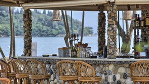 A bar with stone pillars, wicker chairs, and a view of the water. - Molfetta Beach Hotel (Corfu)