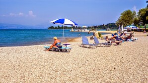 A pebbled beach with people sunbathing under umbrellas, a clear sky, and a calm sea. - Molfetta Beach Hotel (Corfu)