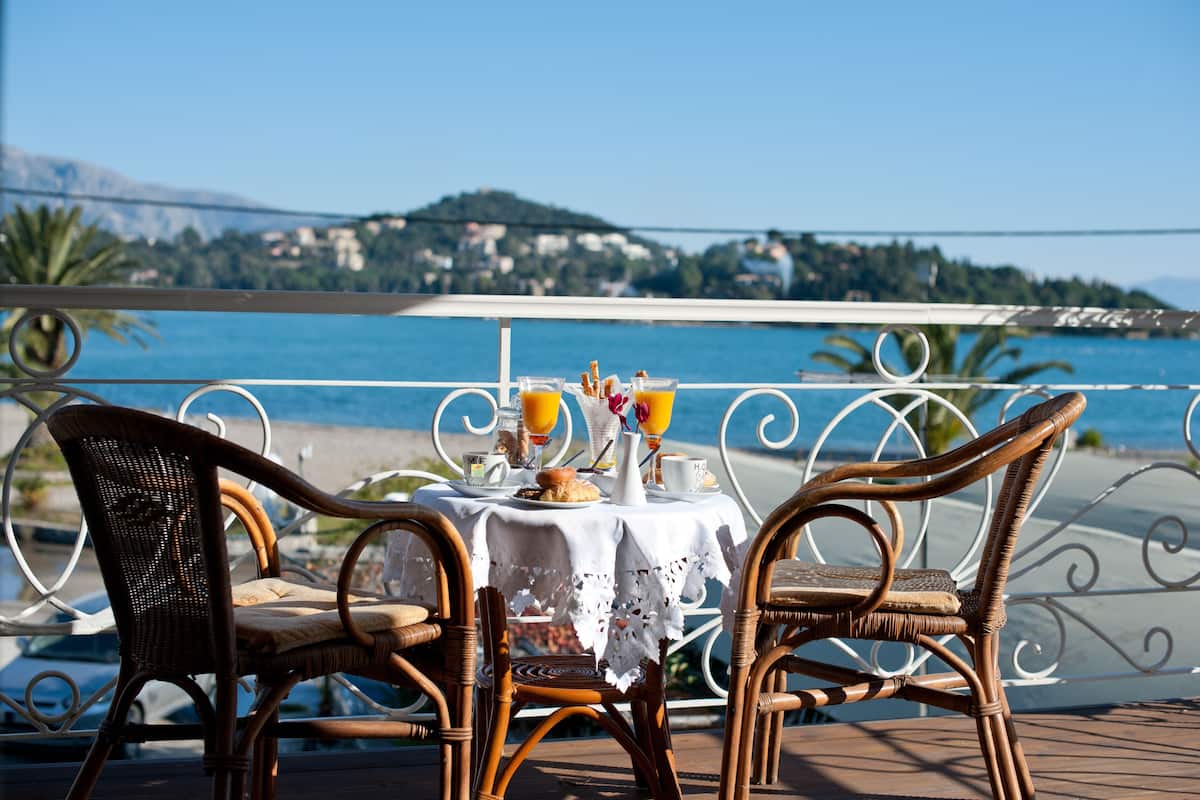A table with wicker chairs and a white tablecloth, overlooking a coastal view with a bridge and hills in the background.
