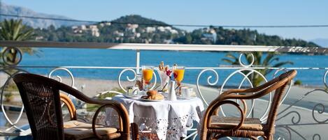 A table with wicker chairs and a white tablecloth, overlooking a coastal view with a bridge and hills in the background.