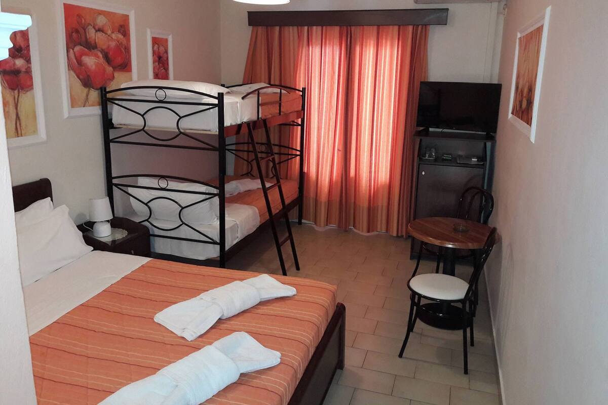 A hotel room with a bunk bed, a single bed, a desk with a chair, a television, and artwork on the walls.
