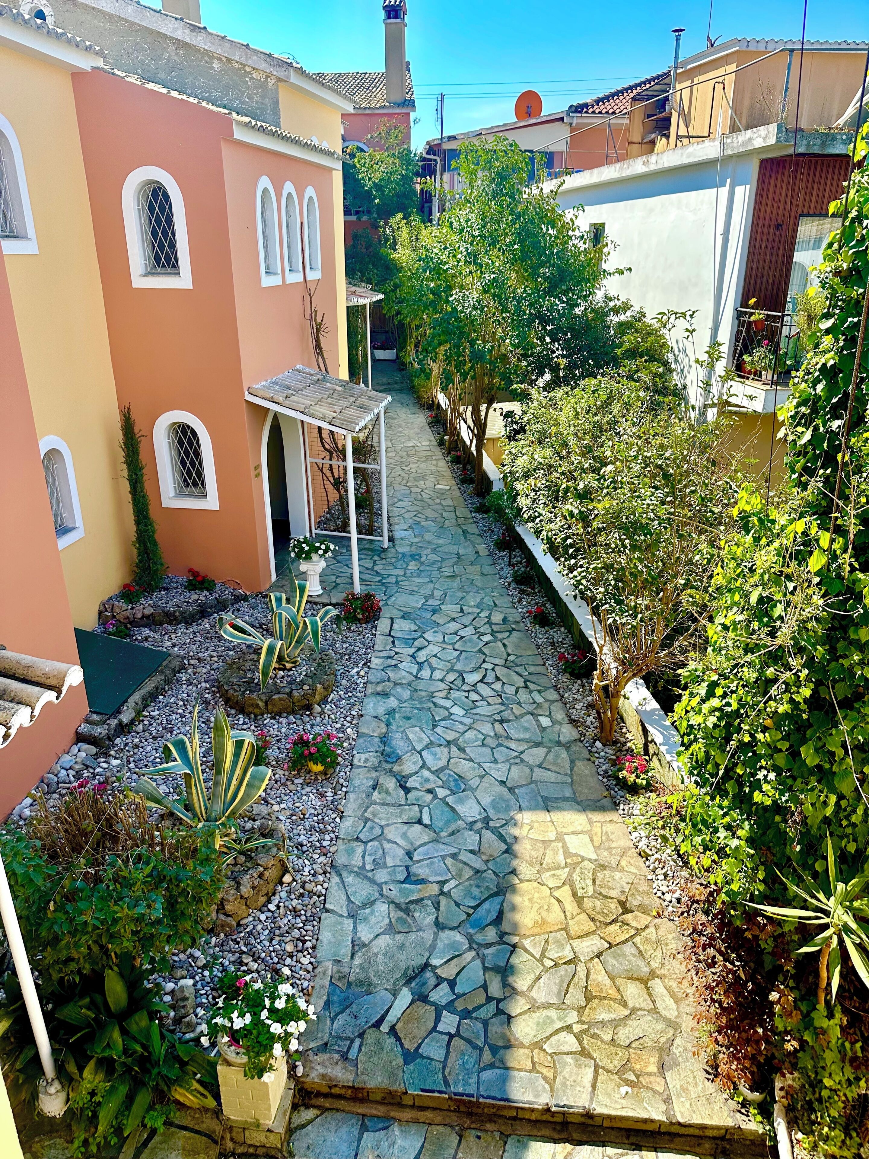 Un étroit sentier de pierre traverse une cour intérieure agrémentée de plantes en pot et d’une pergola.