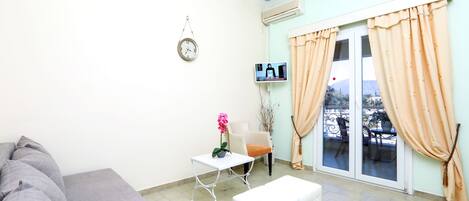 A room with a sofa, a small table with a flower, a chair, a TV, and a window with curtains.