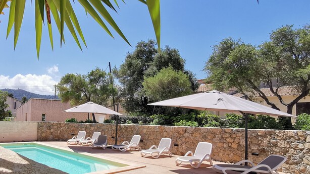 Un espace aménagé au bord de la piscine, avec des chaises longues et des parasols, entouré de murs en pierre et d’arbres.