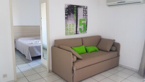 Une petite pièce propre, avec un canapé beige, un coussin vert et un lit recouvert d’une couverture blanche.