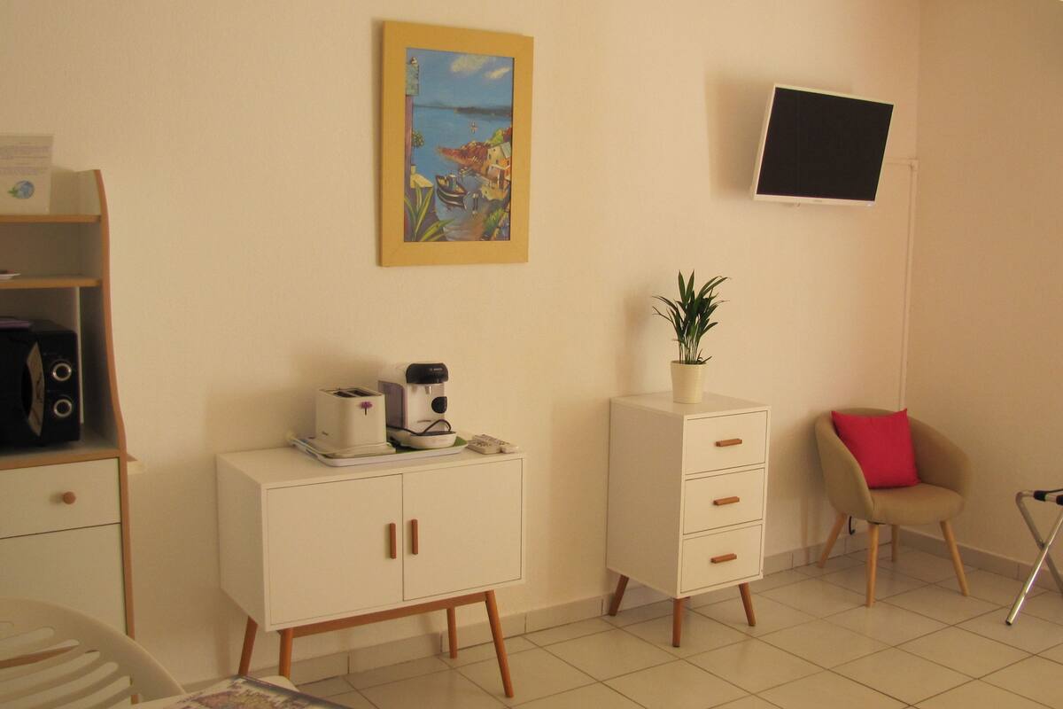 A small, well-lit room with a kitchenette, a single bed, a chair, a desk, and a wall-mounted TV.