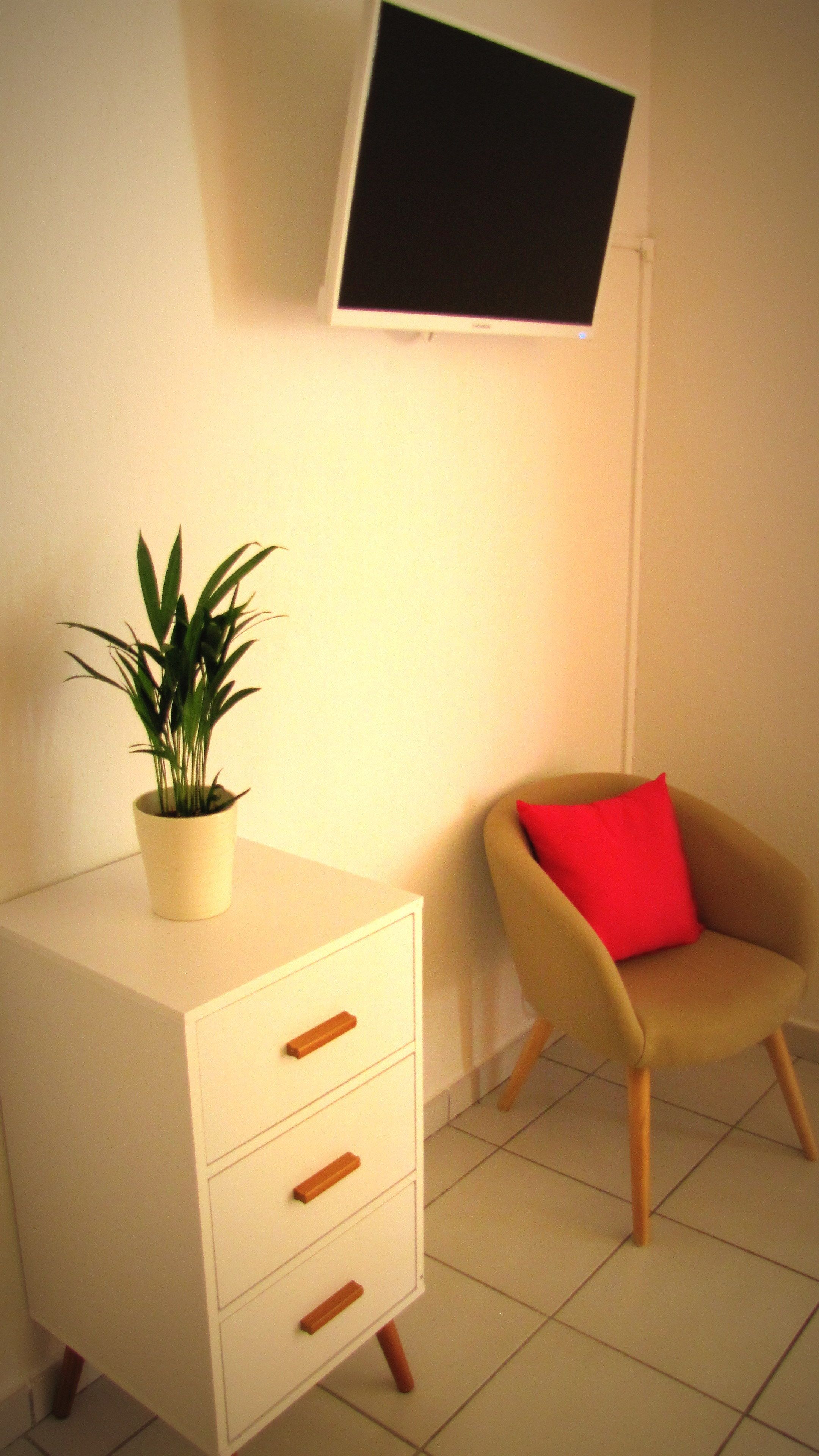 A room with a white dresser, a red chair, a potted plant, and a flat-screen TV mounted on the wall.