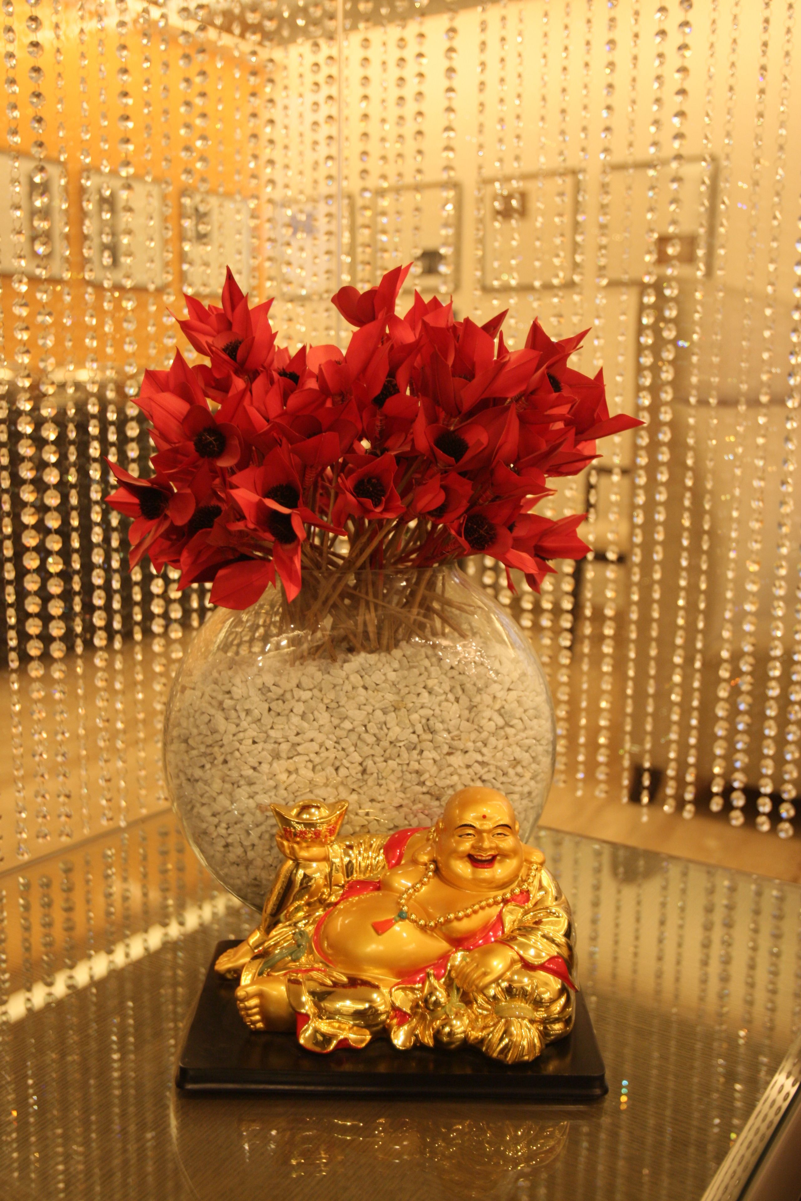 A glass vase with red flowers and a golden Buddha statue on a reflective surface.