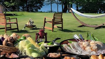 An outdoor dining setup with various food items, a hammock, and chairs in a grassy area with trees and a body of water in the background.