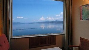 A room with a large window offering a view of a calm body of water and distant mountains. There is a wooden chair and a small table in the room. - Kussharo Prince Hotel (Teshikaga)