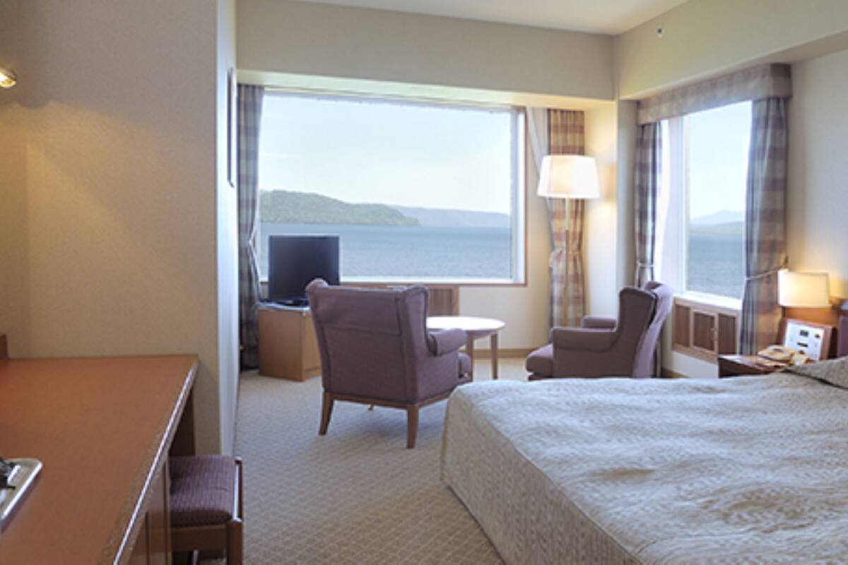 A hotel room with a bed, two armchairs, a small table, a TV, and a window with a view of the sea.