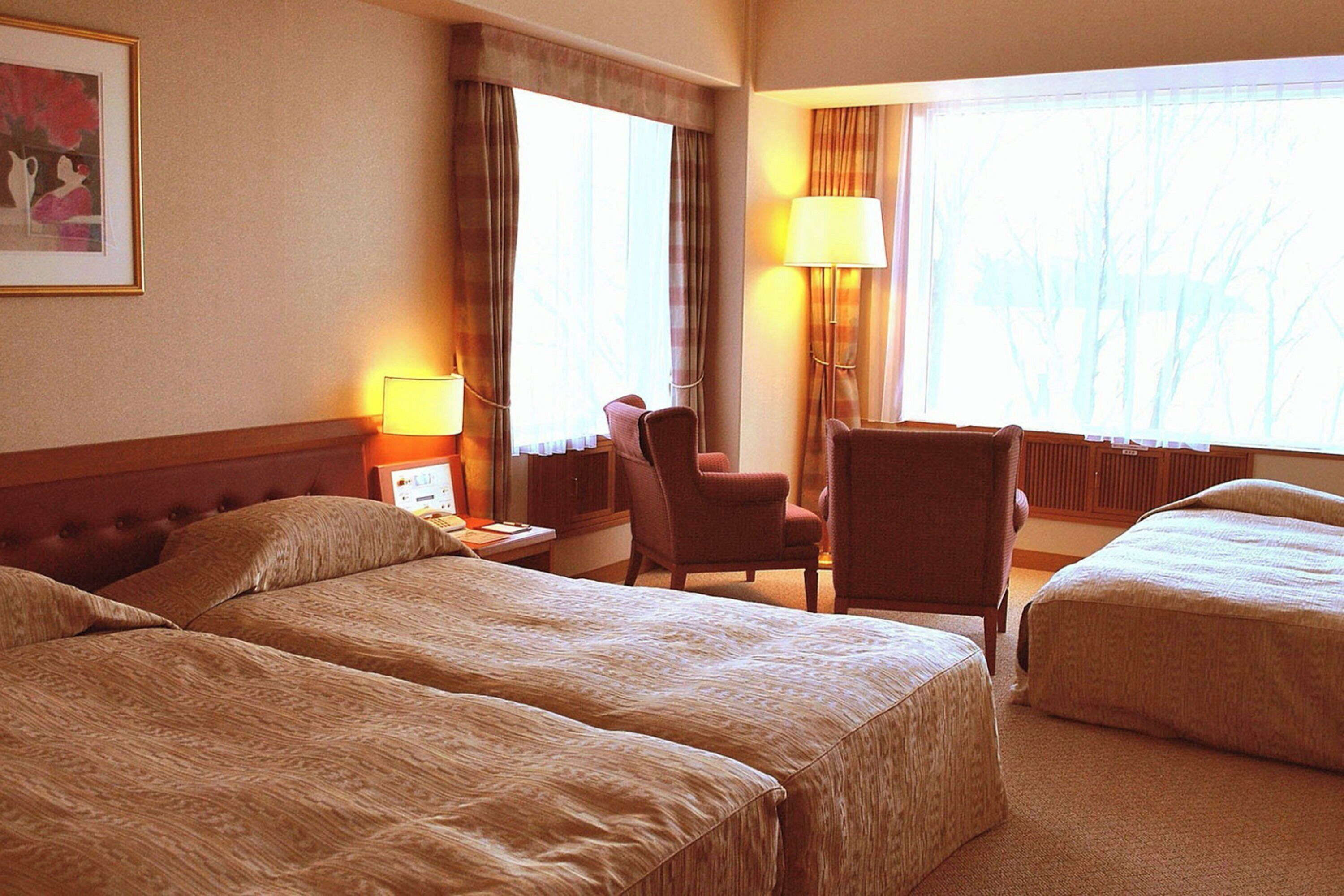A hotel room with two beds, a desk with a lamp, a chair, and a window with curtains.