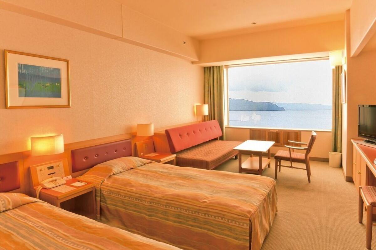 A hotel room with two beds, a sofa, a small table, a TV, and a window with a view of the sea.