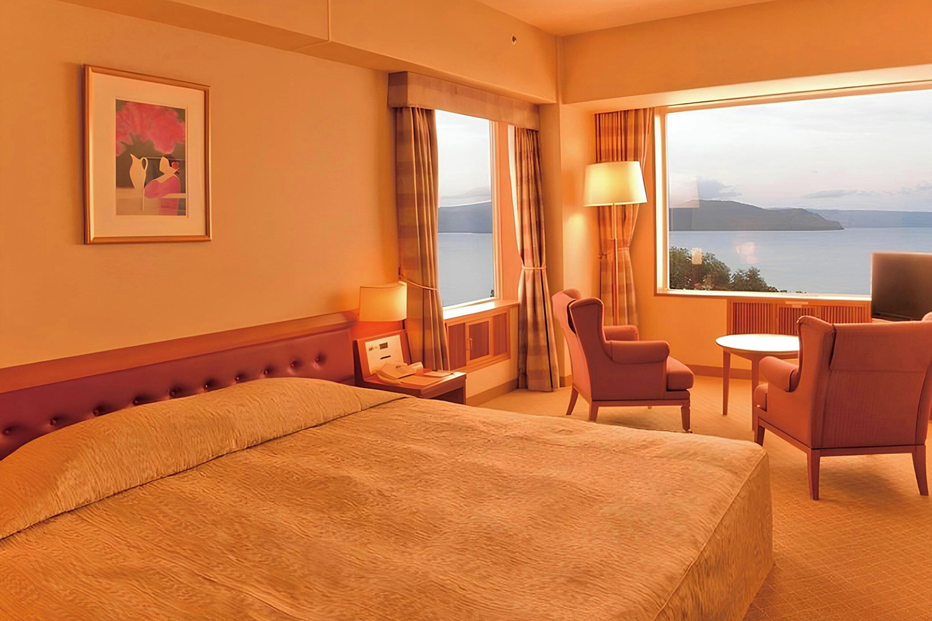 A hotel room with a bed, a desk with a lamp, a chair, a small table, and a view of the water.