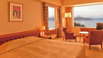 A hotel room with a bed, a desk with a lamp, a chair, a small table, and a view of the water.