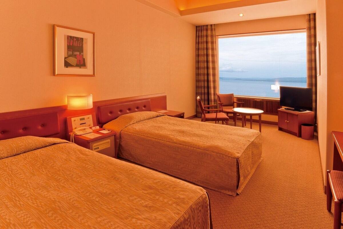 A hotel room with two beds, a desk, a chair, a TV, and a large window with a view.