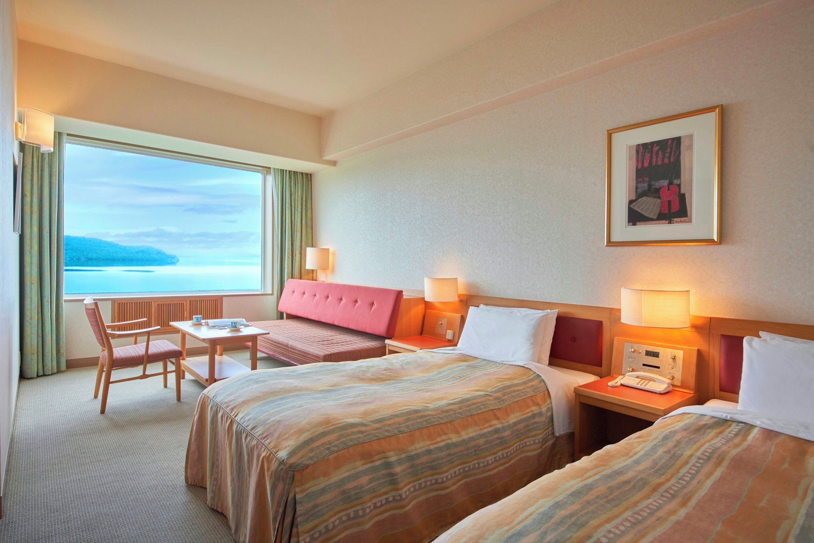 A hotel room with two beds, a desk, a chair, a lamp, a phone, and a window with a view of the sea.