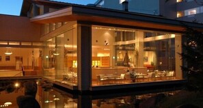 Un hotel moderno con fachada de vidrio, un área de piscina y un espacio de comedor visibles a través de las ventanas.