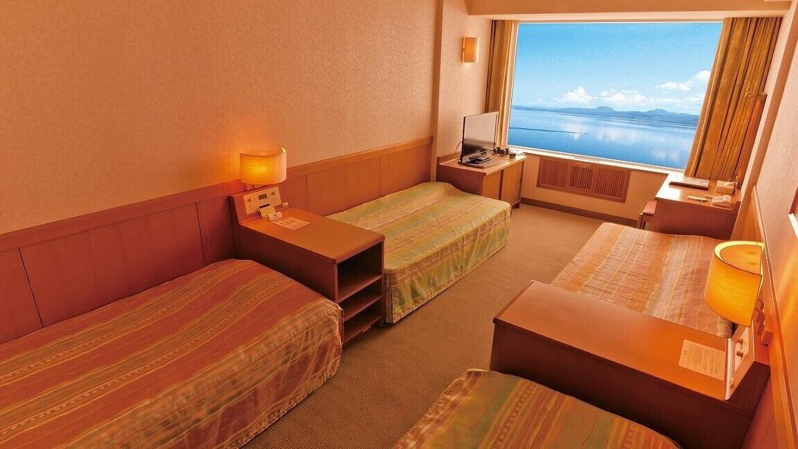 A hotel room with two single beds, a desk, a chair, a lamp, and a window with a view of the sea.