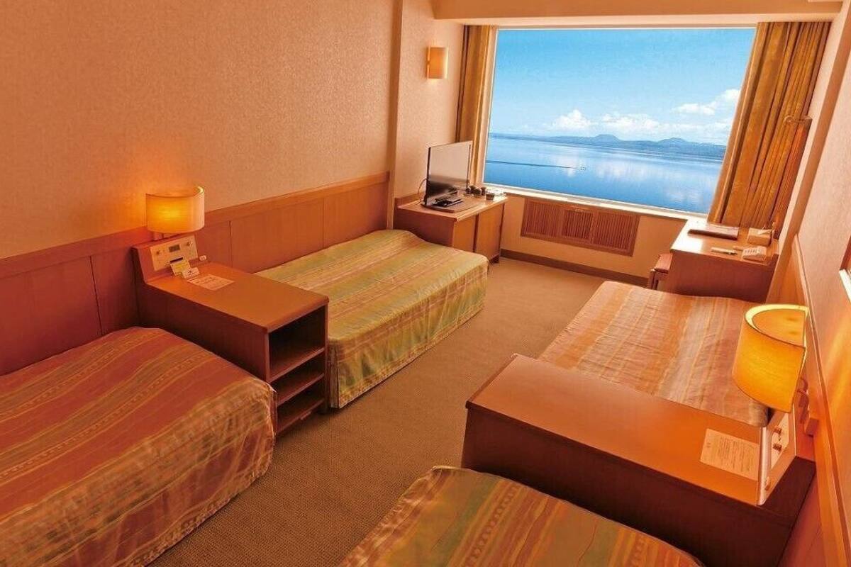 A hotel room with two single beds, a desk, a chair, a lamp, and a window with a view of the sea.