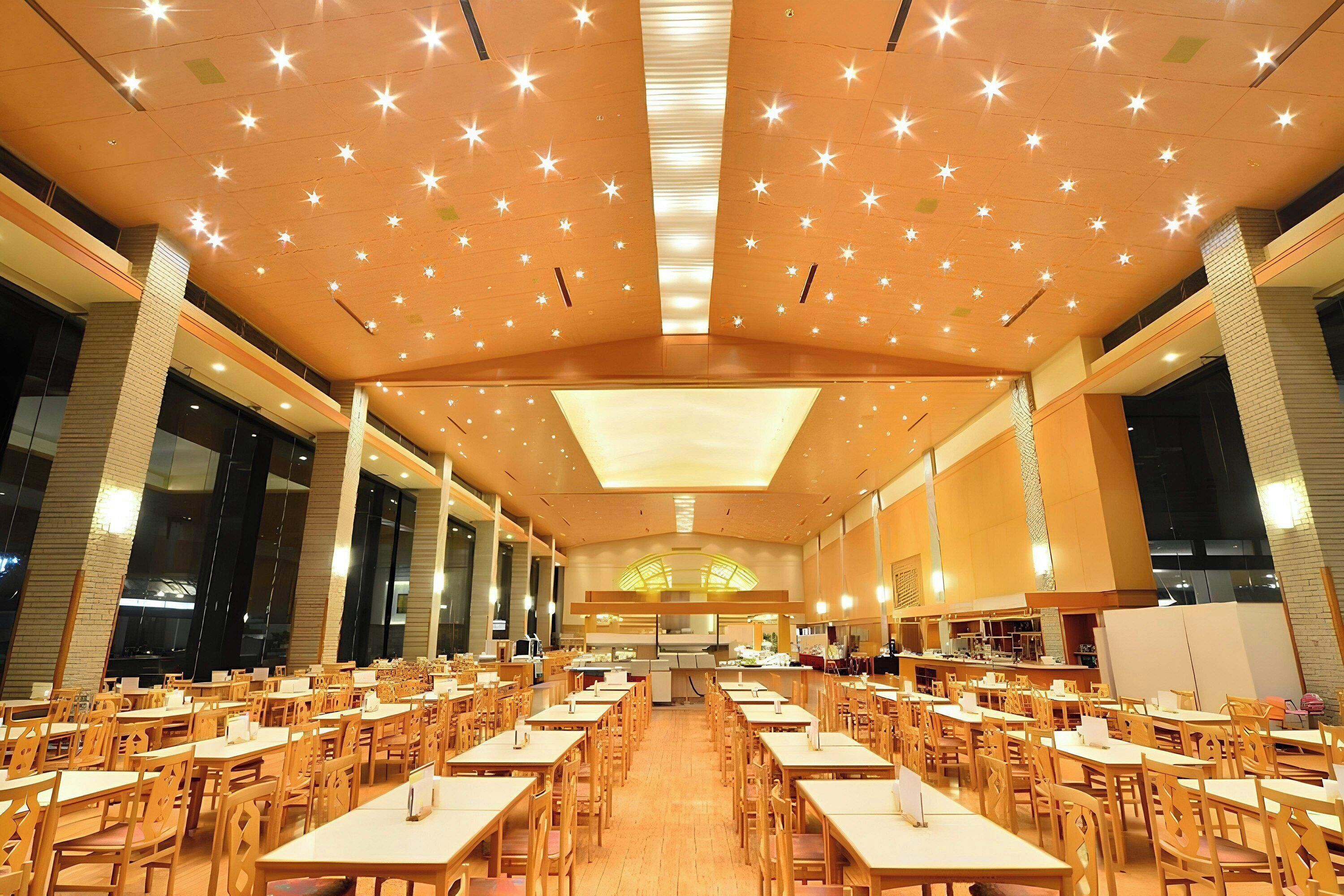 A spacious dining hall with multiple tables and chairs, large windows, and a high ceiling with star-shaped lights.