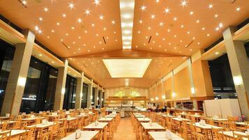 A spacious dining hall with multiple tables and chairs, large windows, and a high ceiling with star-shaped lights.