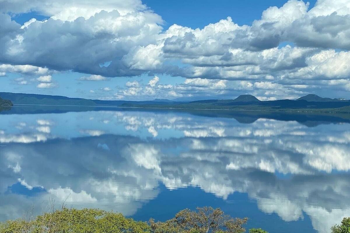 A calm lake with reflections of clouds, surrounded by hills and clear skies.