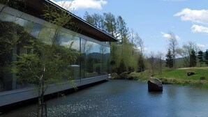 A modern building with a glass facade, a body of water, and surrounding greenery. - Kussharo Prince Hotel (Teshikaga)