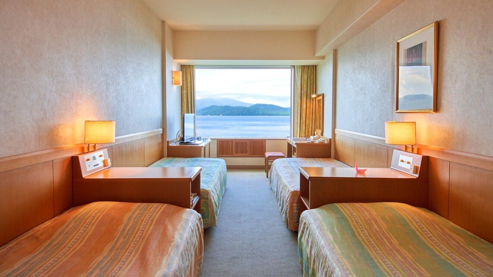 A hotel room with two beds, a desk, a chair, a window with a view of the sea, and a framed picture on the wall.